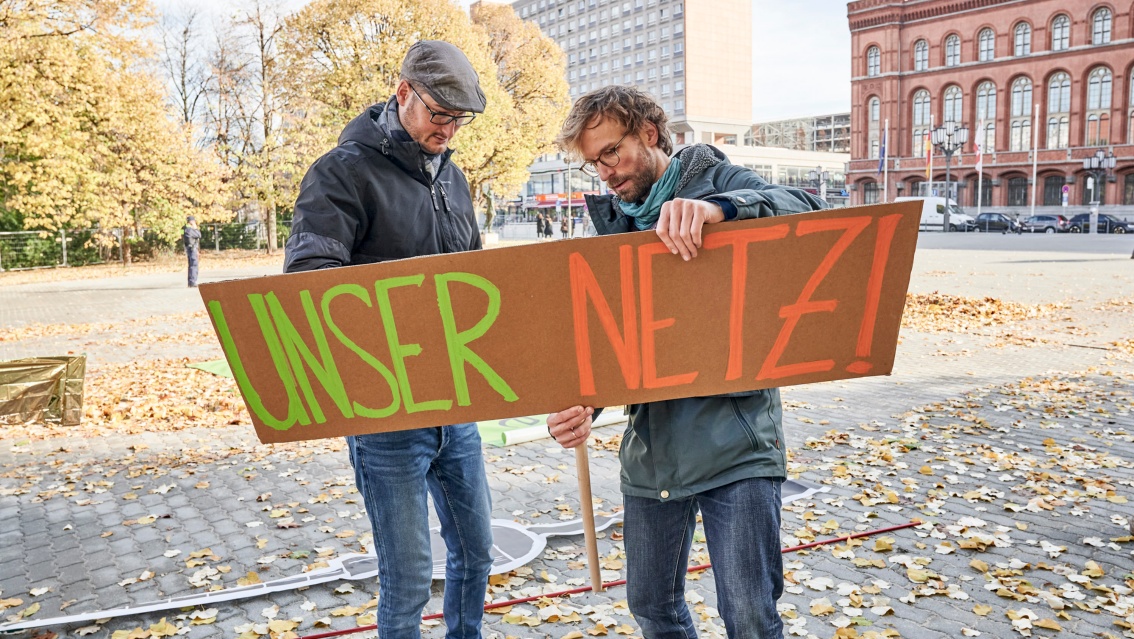 Two young men stand in front of Berlin’s Rotes Rathaus assembling a cardboard sign that displays the slogan “Our grid”.