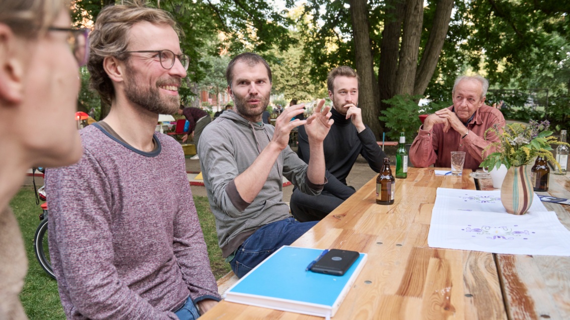 An einem Gartentisch sitzt im Sommer eine Gruppe fünf Menschen und diskutieren miteinander.