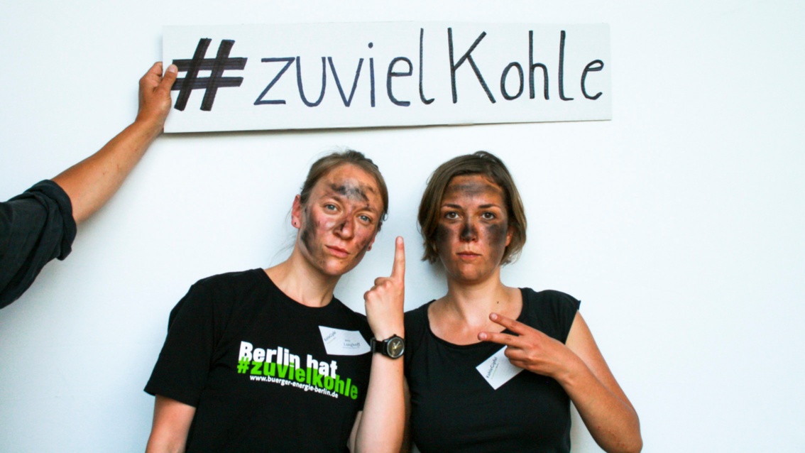 Zwei junge Frauen mit geschwärzten Gesichtern deuten auf ein Schild über sich, darauf steht: Hashtag, zu viel Kohle.