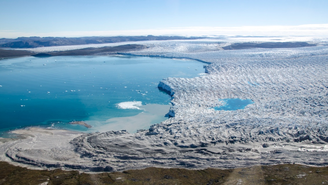 Auf einer Luftaufnahme ist eine große flache Gletscherfläche zu sehen, die in einer geschwungenen Linie an eine Wasserfläche grenzt.