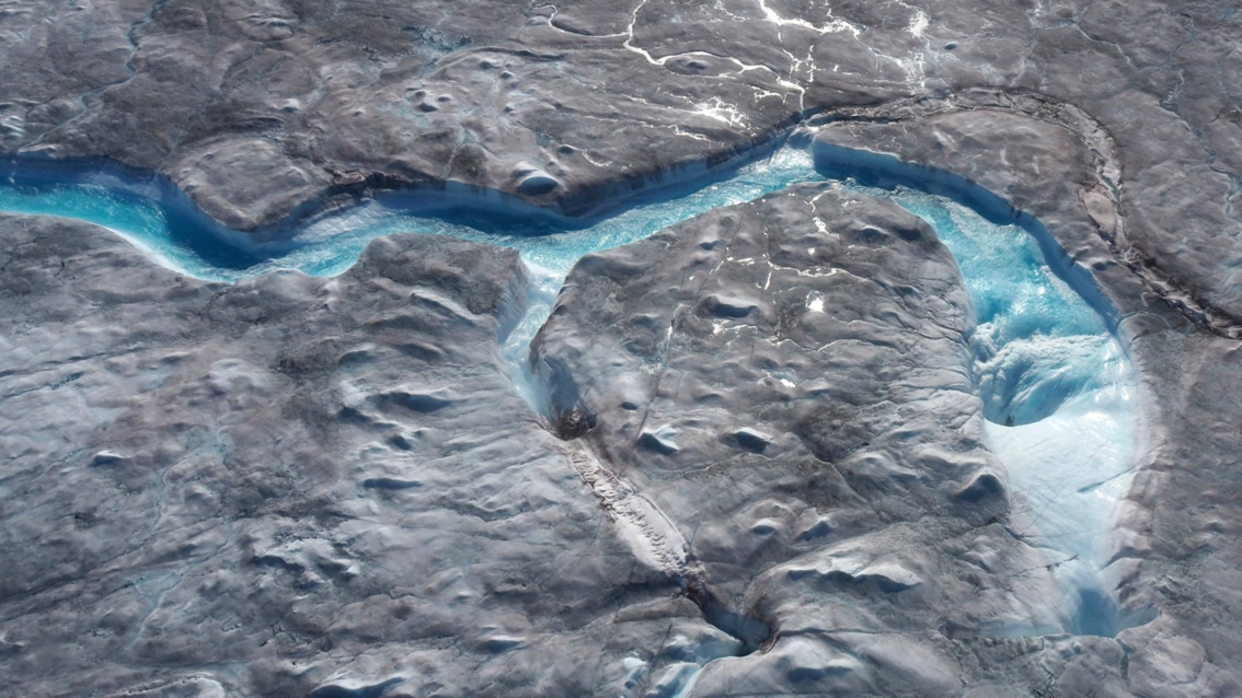 Durch eine braune Eislandschaft mäandert ein Fluss. Das Wasser fließt in Löchern im Eis ab. 