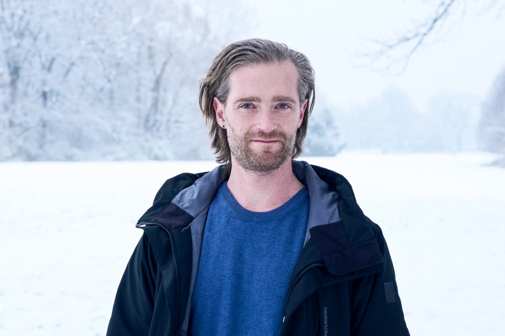 Ein junger Mann mit langem zurückgegelten haaren steht in einer Winterlandschaft