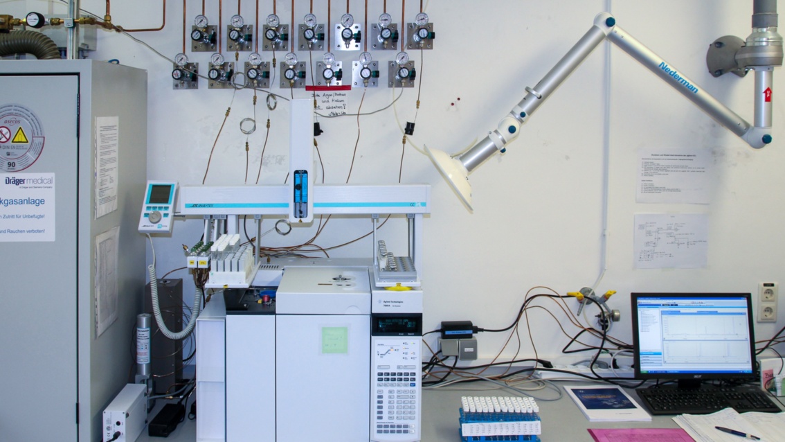 In einem Labor steht ein mikrowellengroßes Gerät auf einem Tisch, an der Wand dahinter Kupferleitungen mit Druckanzeigern.  