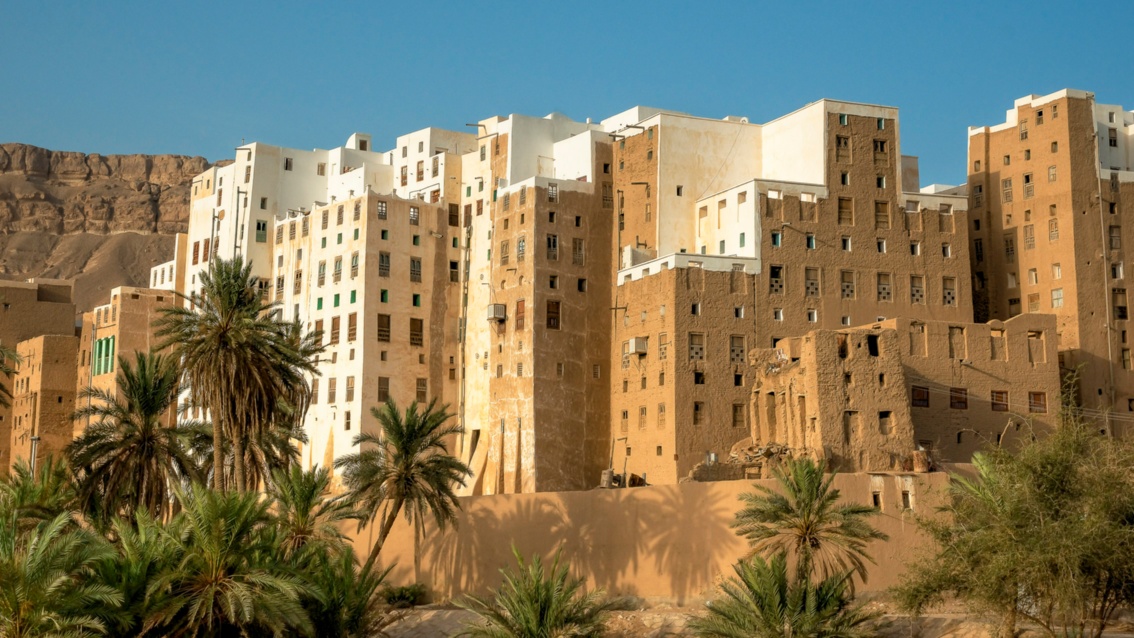 Weiße und lehmfarbene hohe, archaisch anmutende Hochhäuser schließen direkt aneinander an. Dahinter eine blanke Felswand unter strahlend blauem Himmel, im Vordergrund Palmen.