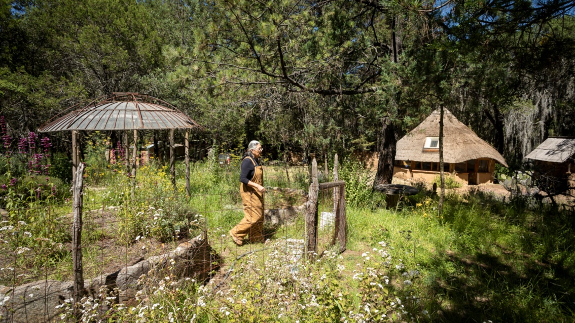 Eine Frau in lehmfarbener Latzhose läuft durch einen grünen wilden Garten, in dem eine runde Laube und ein Haus mit Strohdach zu sehen sind.