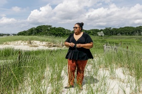Eine dunkelhäutige Frau steht in einem mit Gras bewachsenen Küstenstreifen und schaut Richtung Meer.
