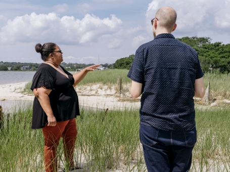 Eine dunkelhäutige Frau steht mit einem weißhäutigen Mann in einem mit Gras bewachsenen Küstenstreifen deutet mit dem Finger Richtung Land.