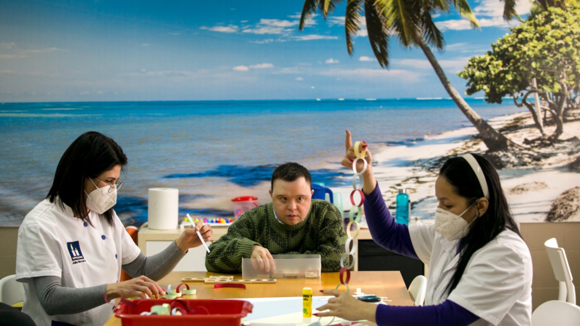 Ein Mensch mit Behinderung sitzt mit zwei Pflegerinnen des Centro  an einem Tisch, hinter ihm eine Fototapete mit Karibikstrand.