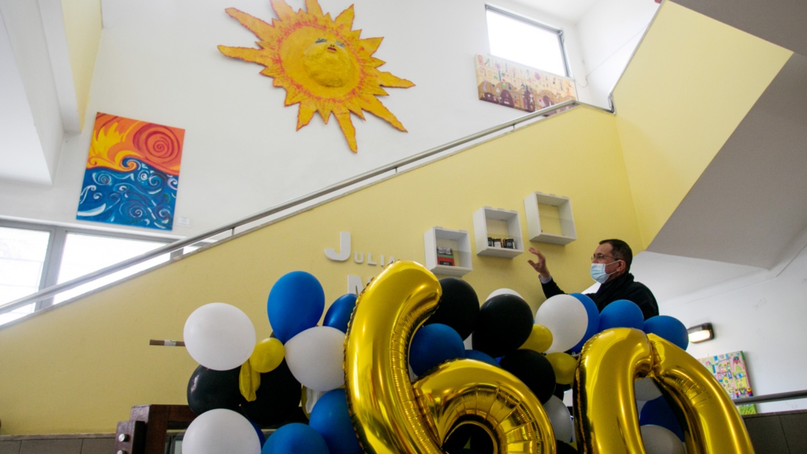 A huge papier-mâché sun hangs in a staircase. Further down, a man stands behind a garland of balloons.