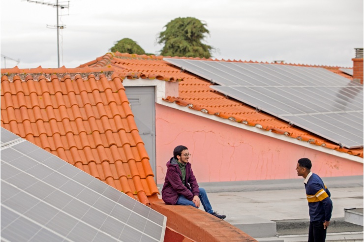 In the midst of a rooftop landscape with PV systems, two young men stand talking happily.