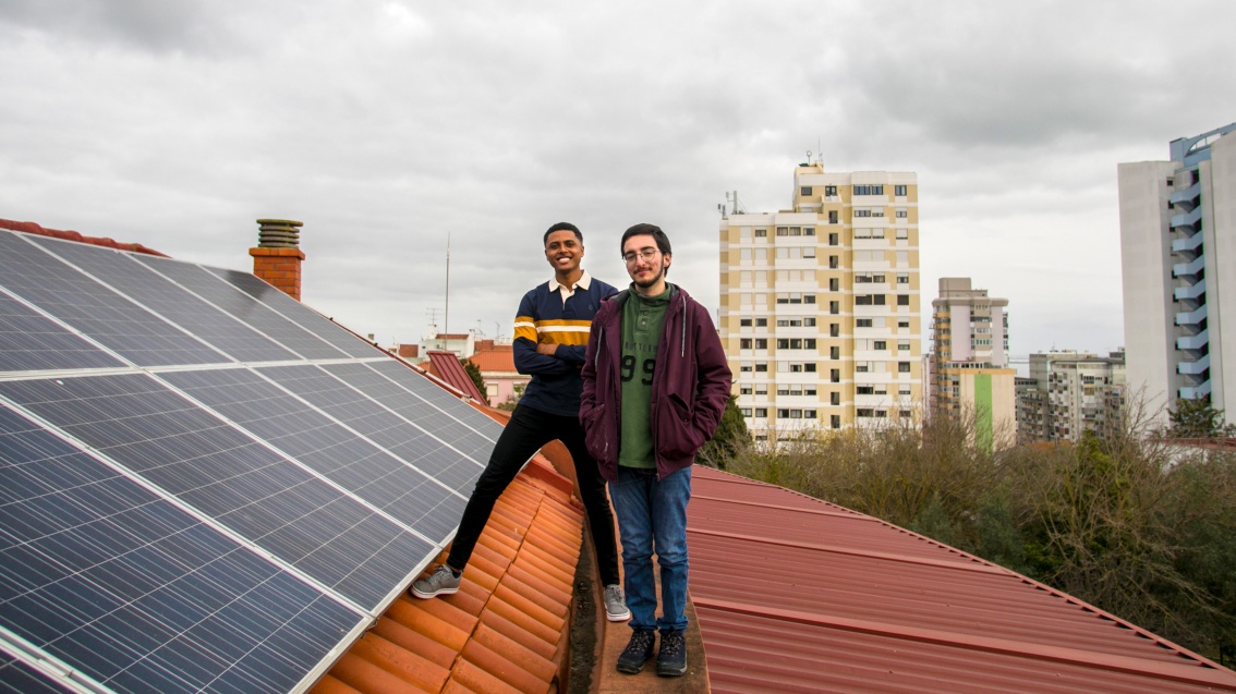 Two young men with friendly smiles pose next to photovoltaic modules on a roof in Lisbon.
