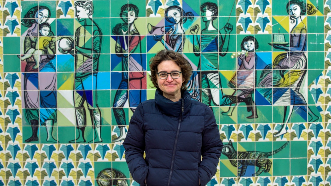 Ana Rita Antunes steht vor einem bunten Wandmosaik und blickt lächelnd in die Kamera.