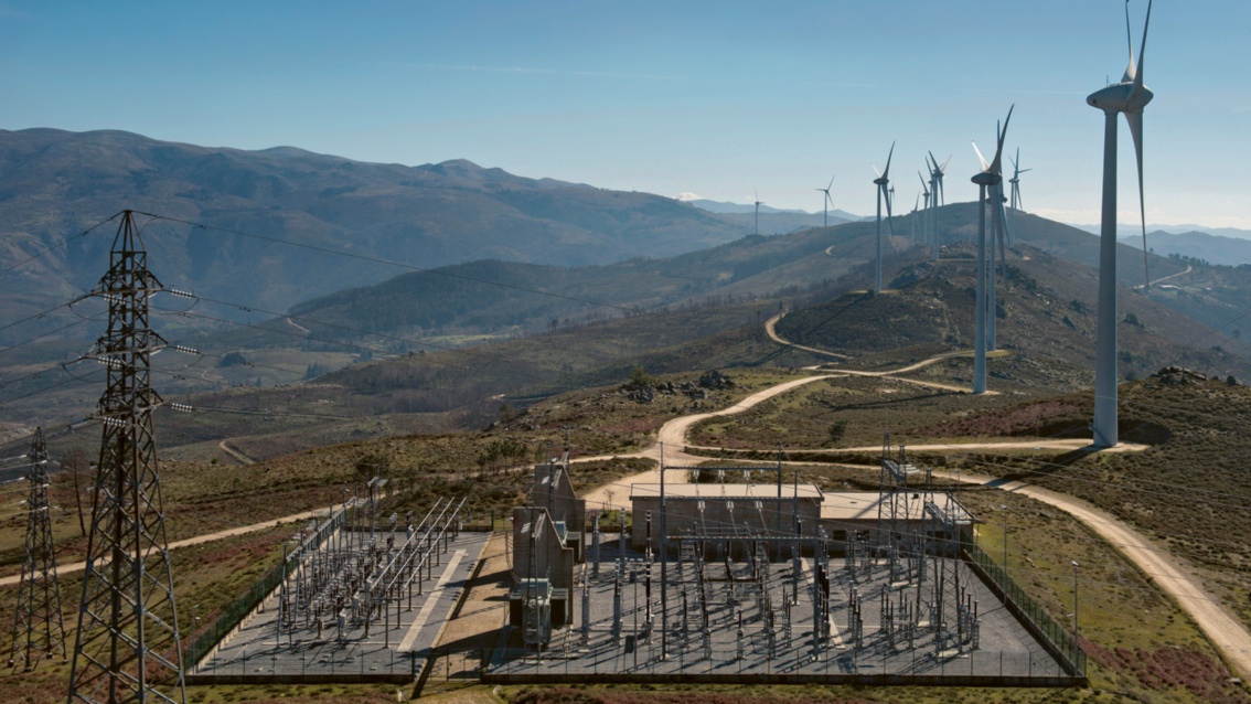 We see a substation in a hilly landscape. A row of wind turbines stretches into the distance over the hills.