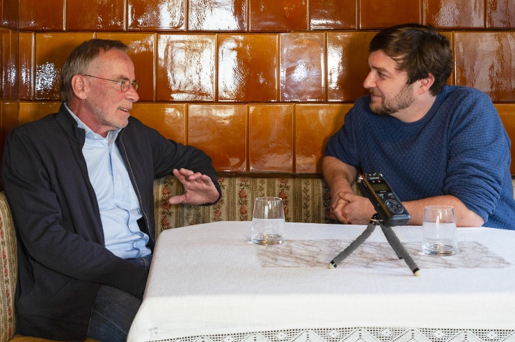 Wolf Dieter Drescher und Sebastian Sladek sitzen vor einem Kachelofen an einem weiß gedeckten Tisch, vor ihnen ein Aufnahmegerät.