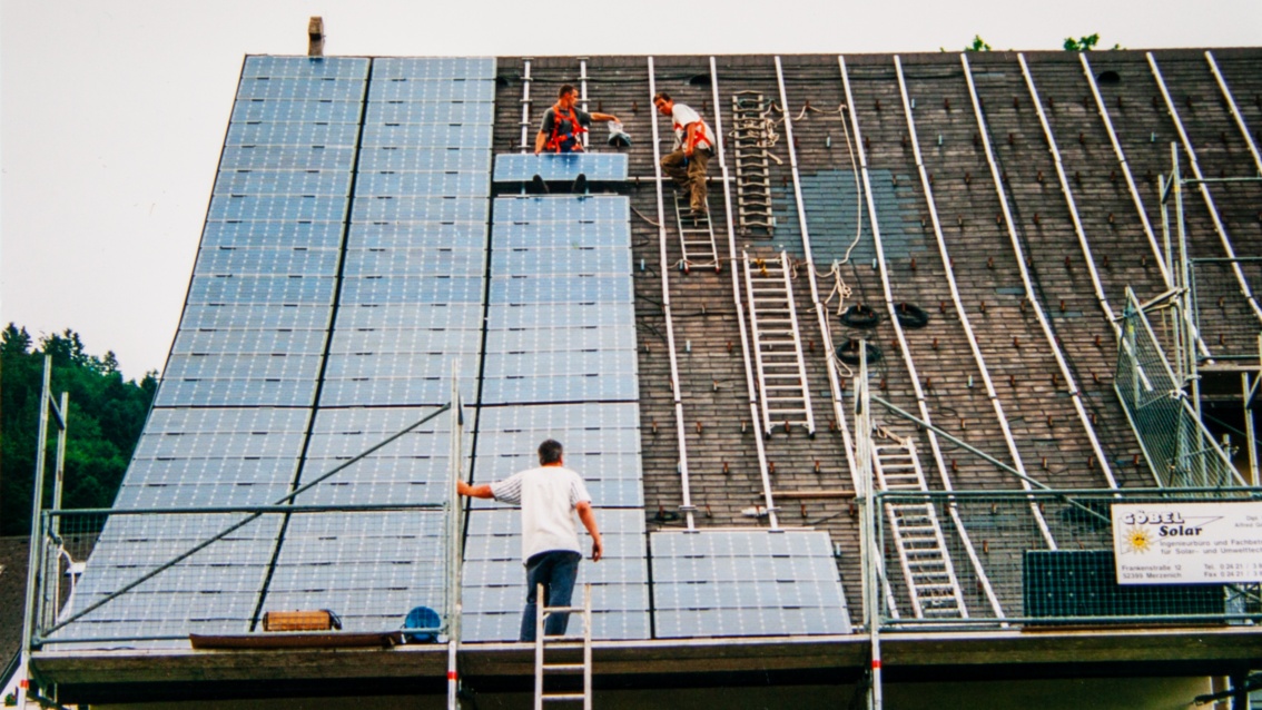 Ein historisches Foto zeigt Arbeiter auf einem steilen großen Dach, die PV-Module installieren.