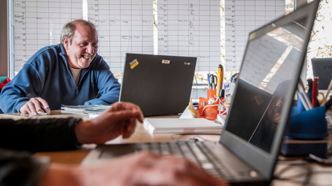 Bei der Arbeit im Büro: André Bechtold sitzt lachend an seinem Notebook. Im Vordergrund arbeitet eine weitere Person am Notebook. 