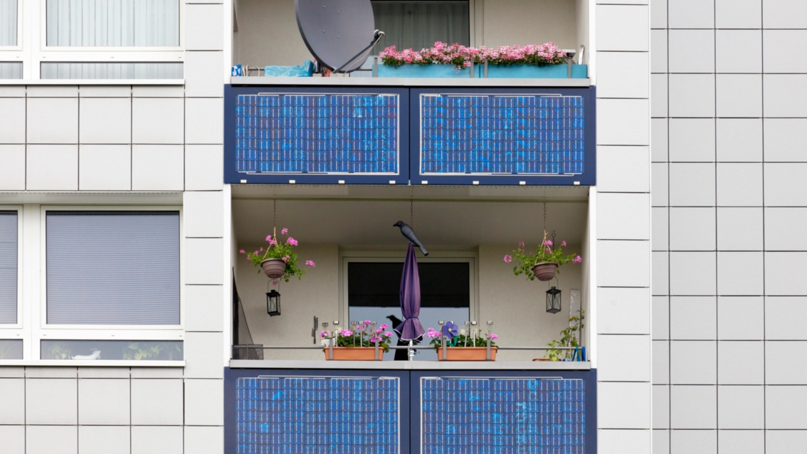 Eine weiße Hausfassade mit zwei Balkons, an deren Brüstungen Solarpanels installiert sind.
