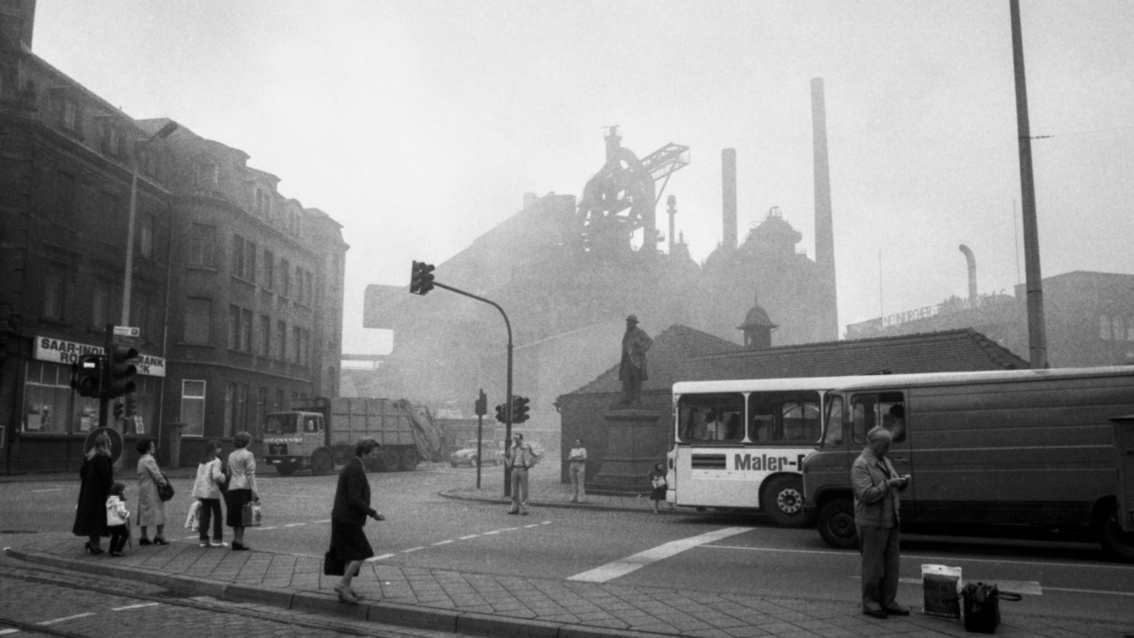 Schwarzweißfoto: Straßenszene mit wartenden Fahrzeugen an einer Fußgängerampel, Smog behindert den Blick auf ein im Hintergrund schemenhaft aufragendes Industriewerk.