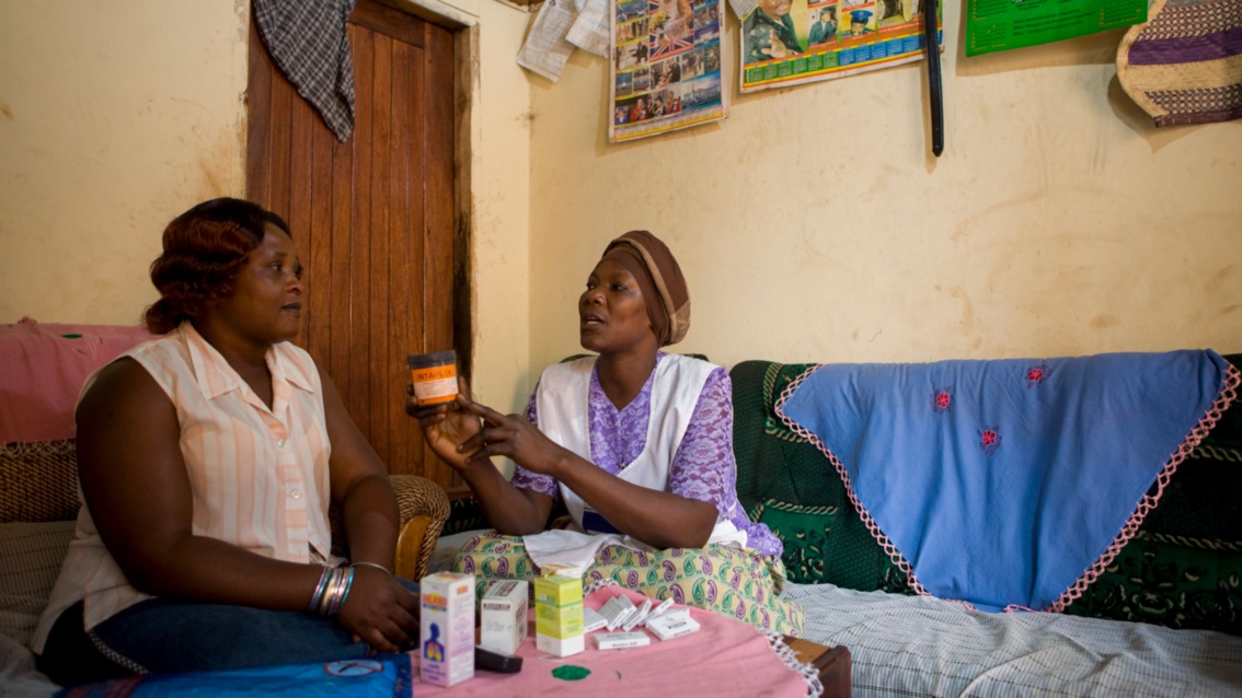 Eine Frau mit schwarzer Hautfarbe sitzt mit einer anderen Frau an einem Tisch und zeigt ihr eine Medikamentendose.
