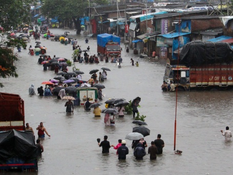 In einer überfluteten Straße laufen Menschen mit Regenschirmen durch das hüfthoch stehende Wasser, Lastwagen stehen quer 