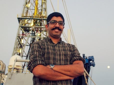 Ein junger Mann mit schwarzem Schnurrbart und Brille steht mit verschränken Armen an Deck eines Schiffes vor einem Bohrturm und lächelt in die Kamera.