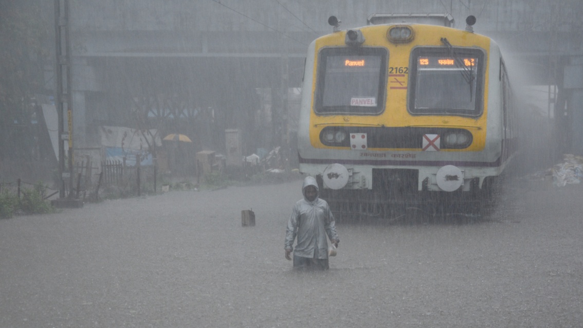 Ein Zug steht im Monsunregen, das Gleisbett ist komplett überflutet. Davor steht ein Mann knietief im Wasser.
