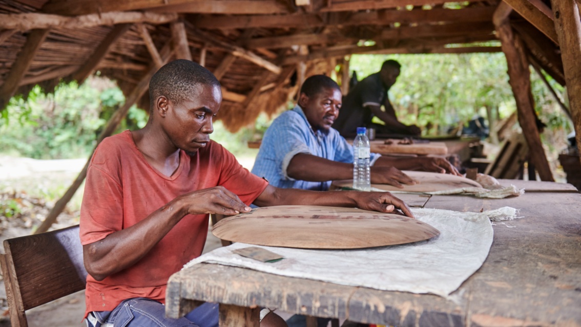 Drei Männer sitzen an einem langen Tischen und bearbeiten runde Holzplatten mit Schleifpapier. 