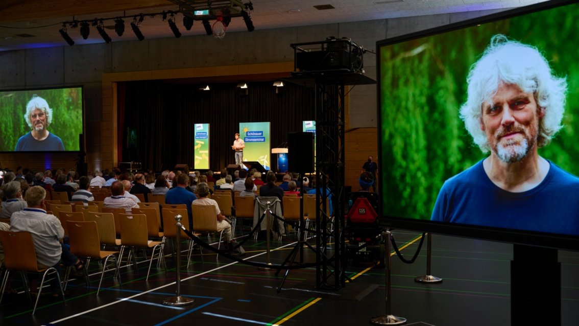 Auf großen Monitoren im Zuschauerraum ist ein Bild von Jochen Stay im Grünen projiziert.