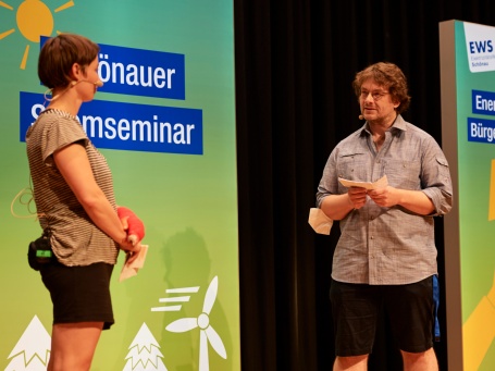 Die beiden Vortragenden stehen in sommerlichen Shorts auf der Bühne und sprechen miteinander.