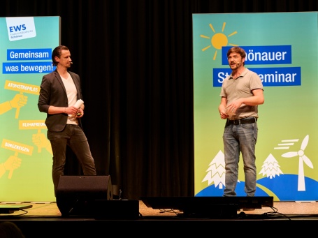 Auf einer Bühne stehen zwei Männer vor den bunten Stelen der Veranstaltung Schönauer Stromseminar.