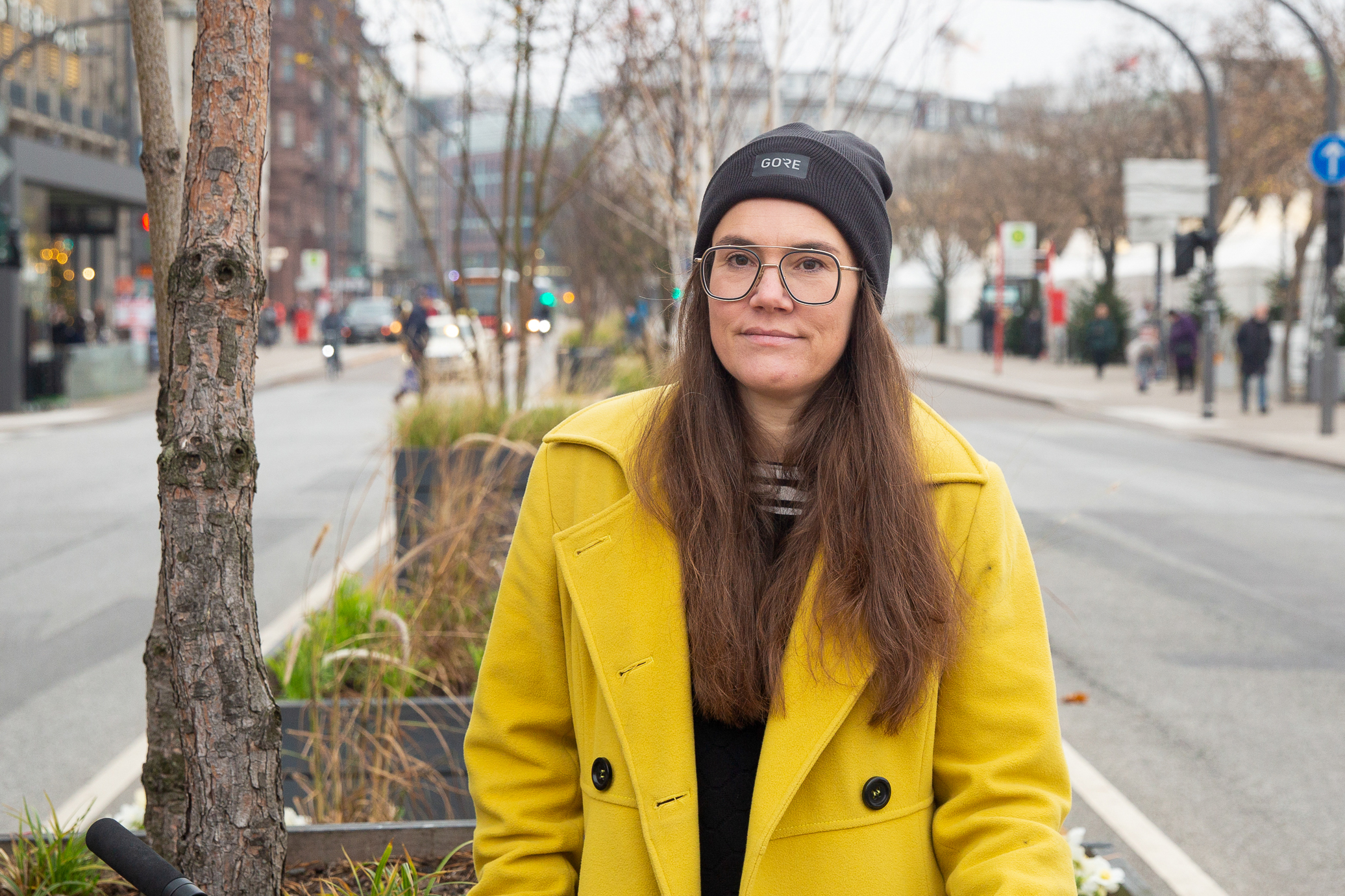 Auf einer städtischen Verkehrsinsel steht eine Frau mit Mütze und in gelbem Mantel und schaut ernst in die Kamera.