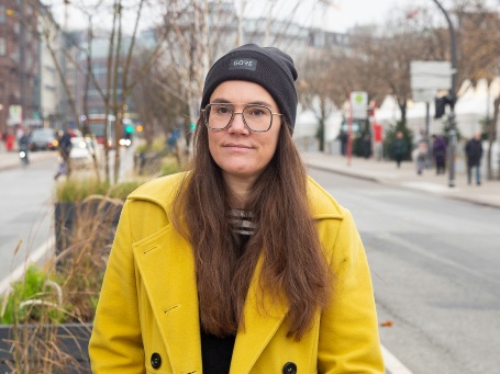 Auf einer städtischen Verkehrsinsel steht eine Frau mit Mütze und in gelbem Mantel und schaut ernst in die Kamera.
