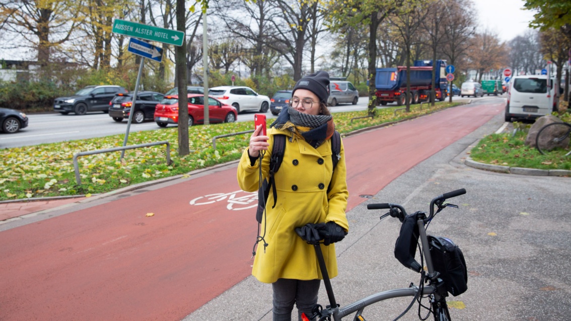Katja Diehl steht mit ihrem Rad auf dem roten Fahrradweg und fotografiert mit ihrem Smartphone.