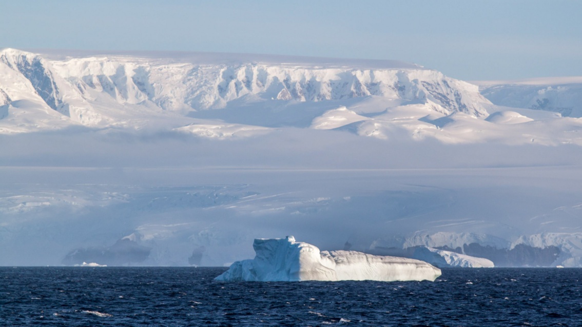 Vor der Küste treibt ein Eisberg im Meer – verglichen mit dem gigantischen Gletscher dahinter wirkt er winzig. 