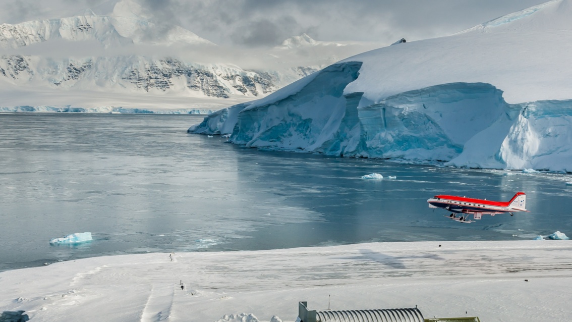 Von rechts schiebt sich ein rot-weiß-blaues Flugzeug in das Gletscher-Panorama, darunter befindet sich eine Landebahn.
