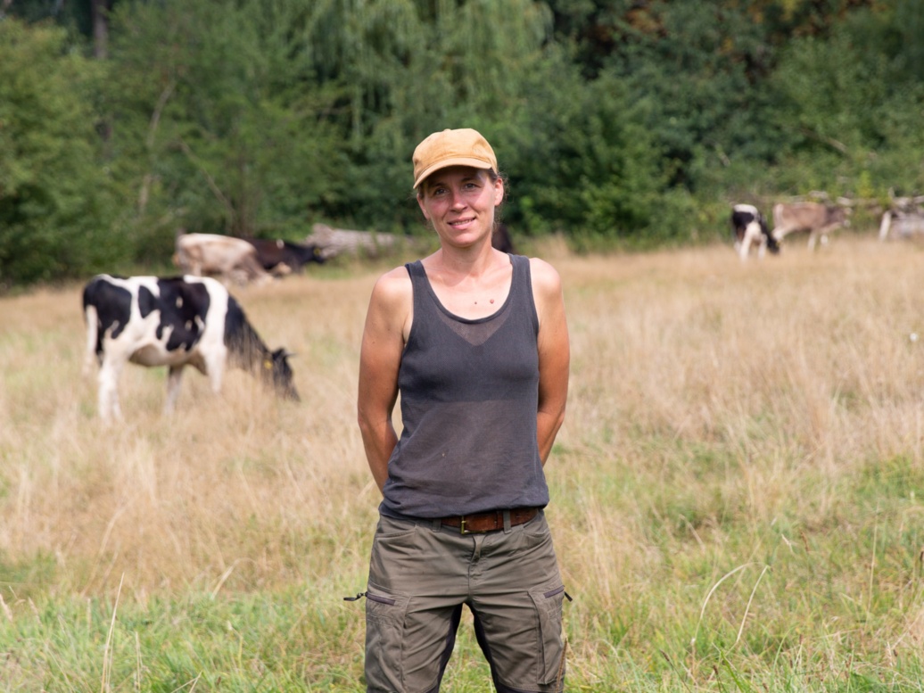 Eine drahtige Frau steht in Arbeitskleidung und mit Käppi auf einer Weide, hinter ihr grasen Kühe.