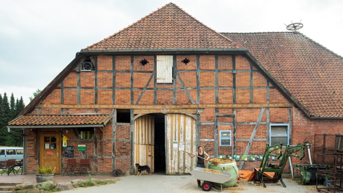 Ein im Fachwerkstil erbautes Bauernhaus in der Totalen: davor schaufelt Annabelle Gérard Heu in eine Schubkarre. 