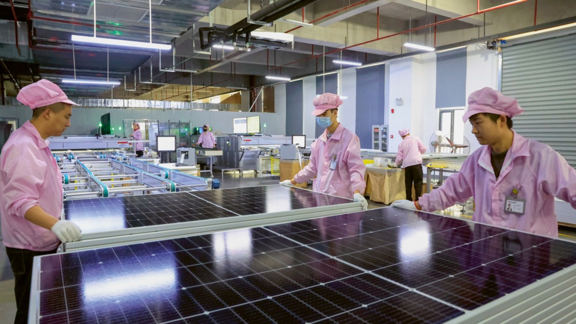 In einer Fertigungshalle betrachten Arbeiter in rosa Schutzkleidung die Oberfläche eines Solarmoduls.