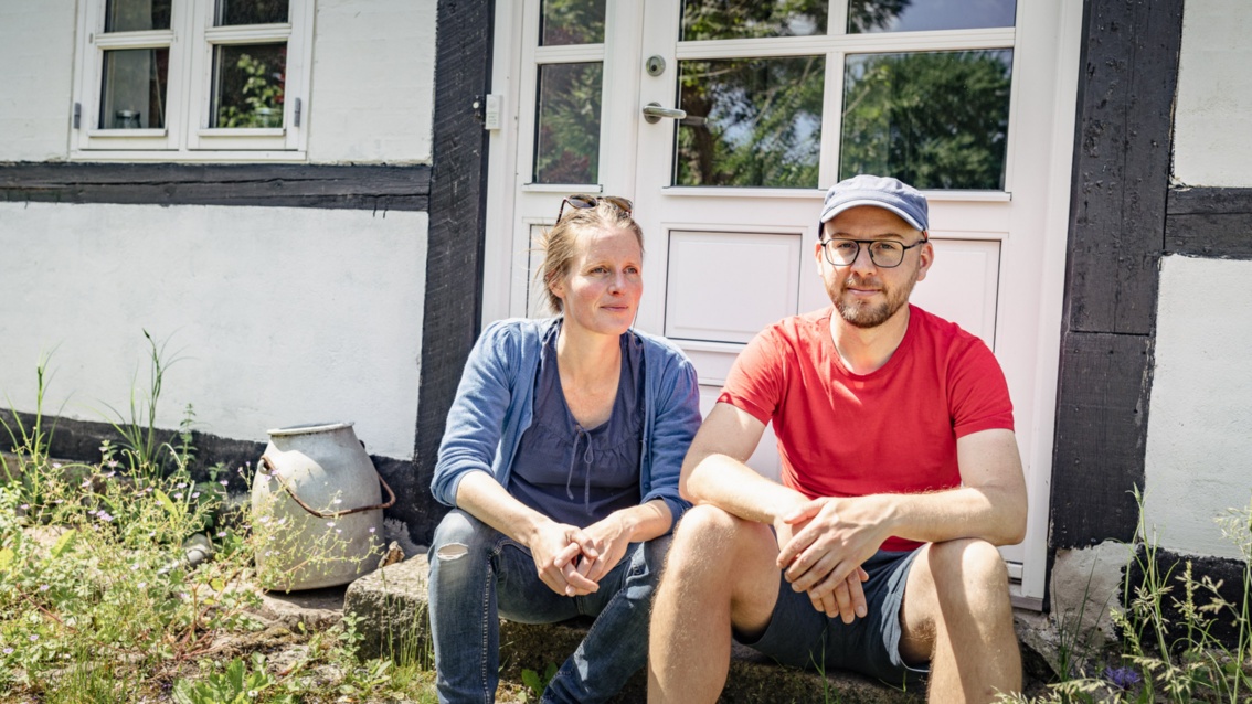 Eine junge Frau und ein junger Mann sitzen auf einer Türschwelle, die zu einem weiß verputzten Fachwerkhaus gehört.
