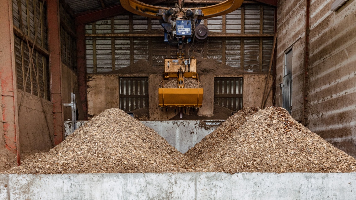 Two piles of shredded wood lie in a small hall - an orange hydraulic grapple hangs above them.