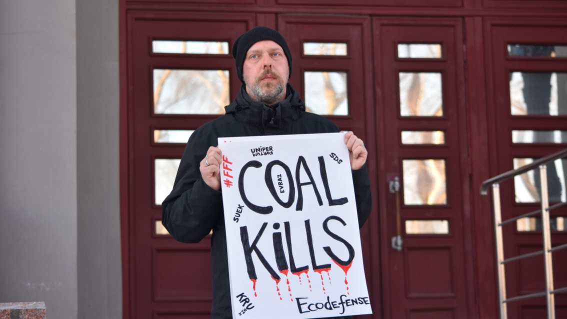 Slivyak steht alleine und mit ernstem Blick vor einem Gebäude, dabei hält er ein Plakat auf dem «Coal kills» steht.