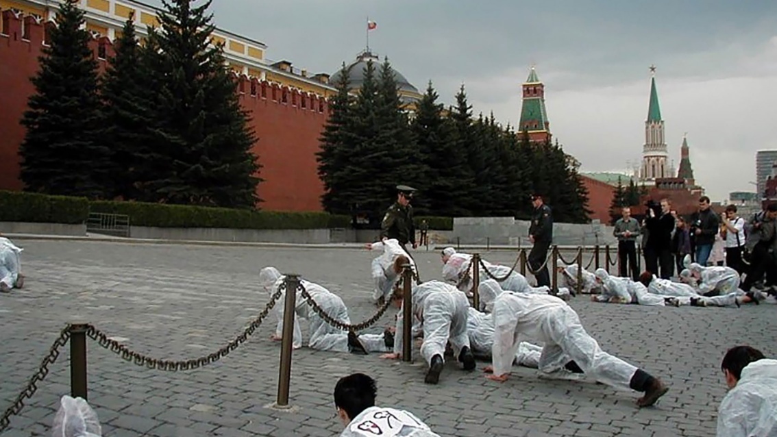 Aktivisten in weißen Schutzanzügen unterqueren kriechend eine Absperrung am Kreml, Soldaten beäugen sie dabei.