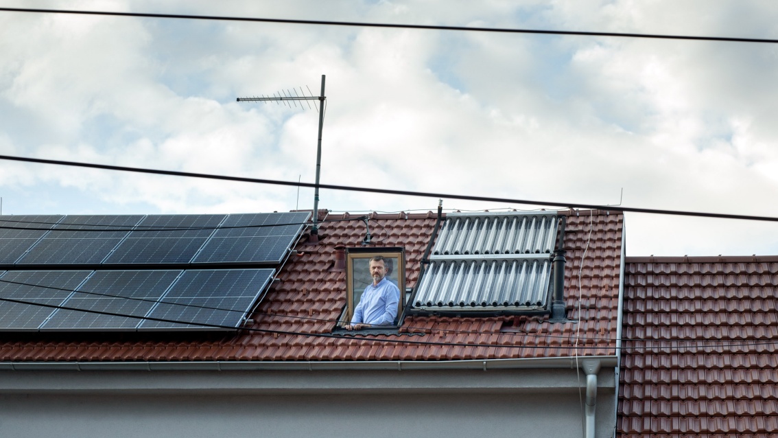 Photovoltaic modules are installed on the roof of a terraced house, and between them a man sticks out of a skylight and looks into the camera.
