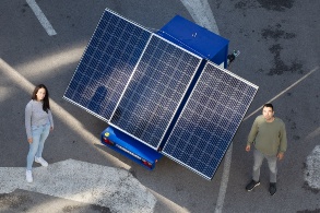 A young man and a young woman stand in front of a solar module mounted on a trailer and look friendly into the camera.
