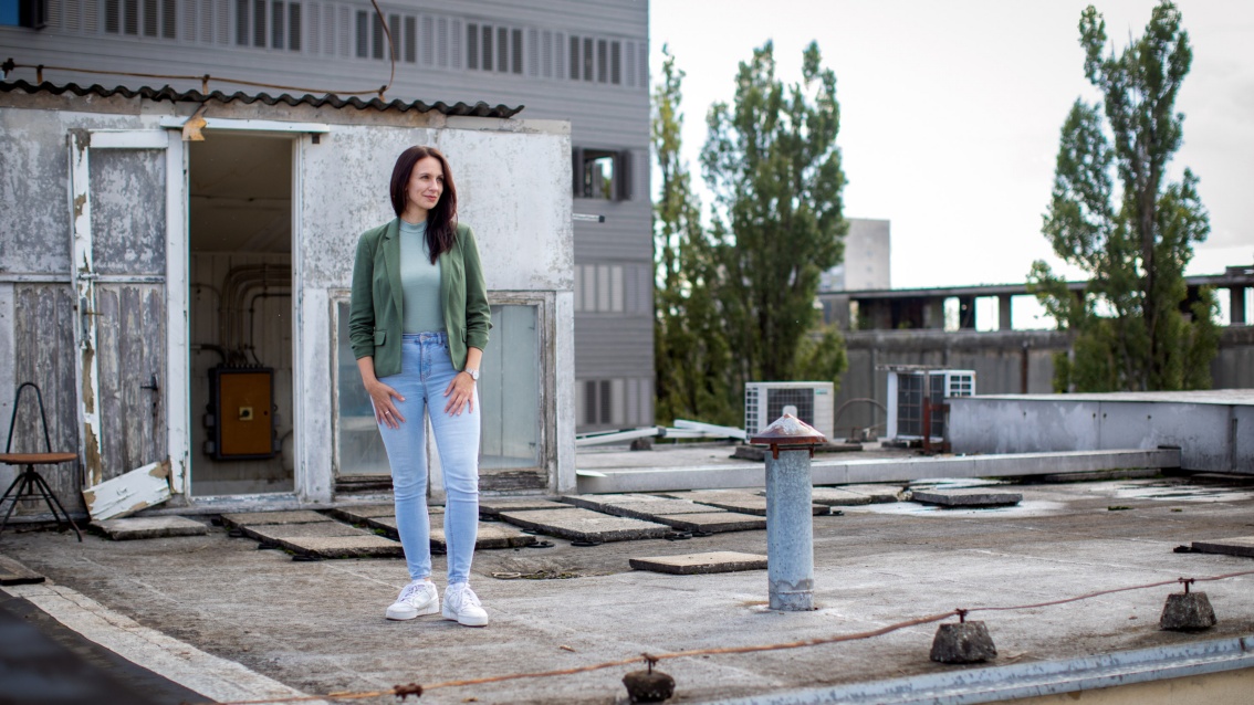 A middle-aged woman in light jeans and a green blazer stands on a flat roof and looks into the distance. 