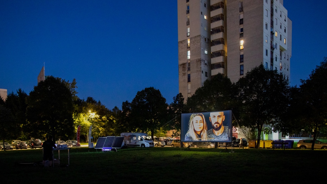 An open-air cinema is set up on the lawn in front of a high-rise residential building, with photovoltaic modules next to the screen.