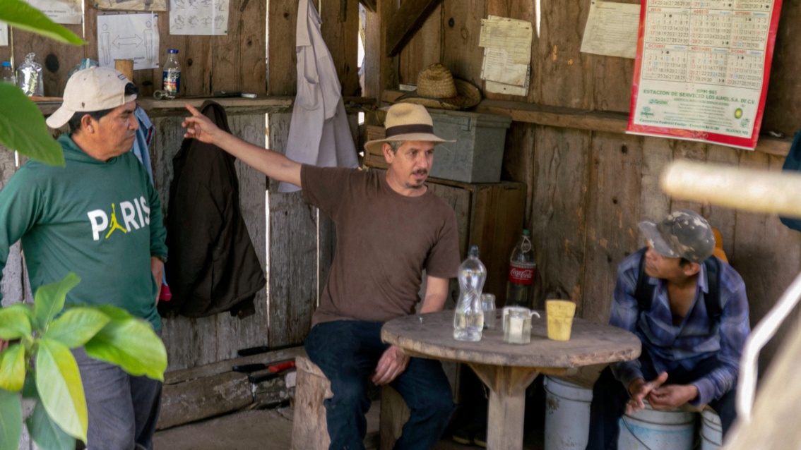 Drei Männer unterhalten sich in einer Holzhütte, auf einem kleinen Tisch stehen Becher und Flaschen. 