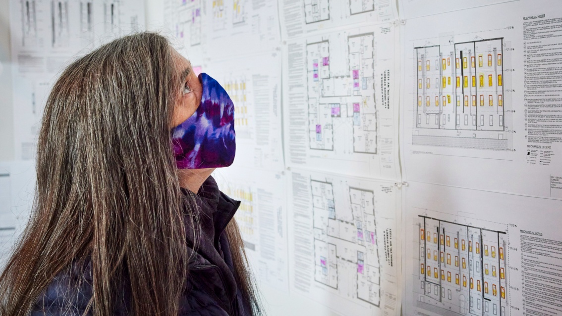 Chris Benedict studiert aufmerksam Baupläne, die an einer Wand hängen. Sie trägt eine Gesichtsmaske mit Batikmuster. 