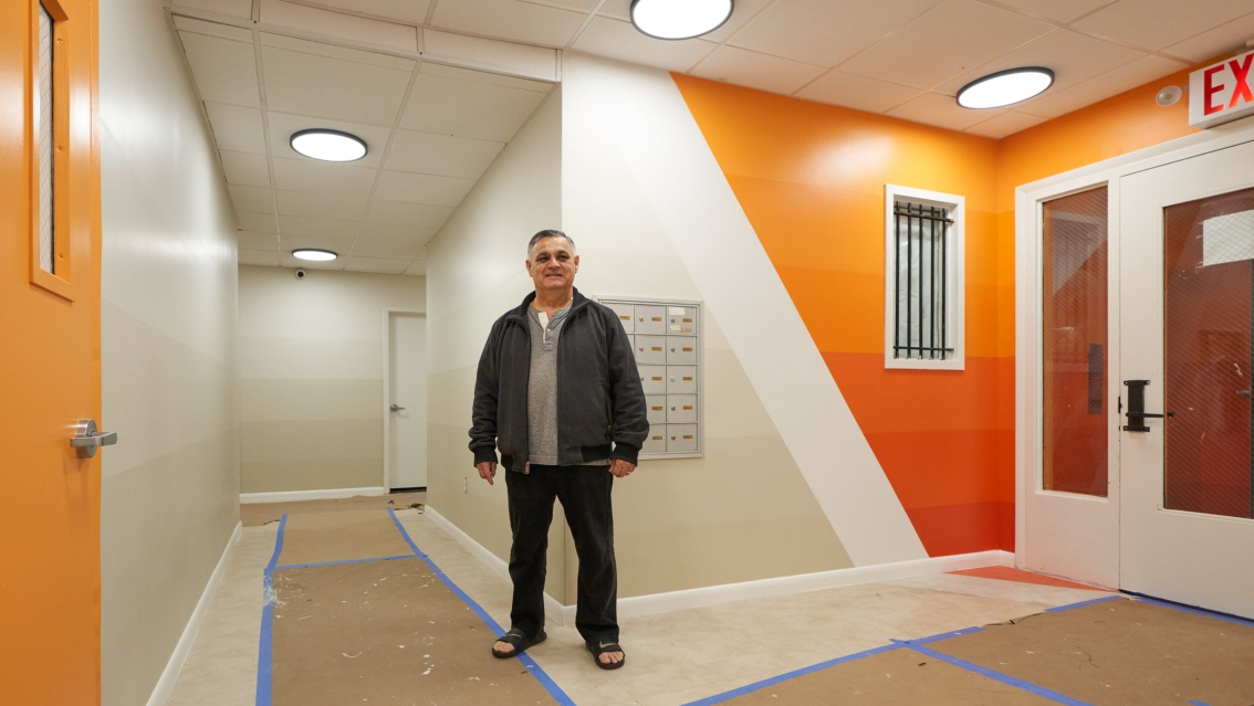 Ein Mann steht lächelnd mit Badelatschen im Hausflur – die Wände sind in abgestuften Orange- und Grautönen gestaltet. 