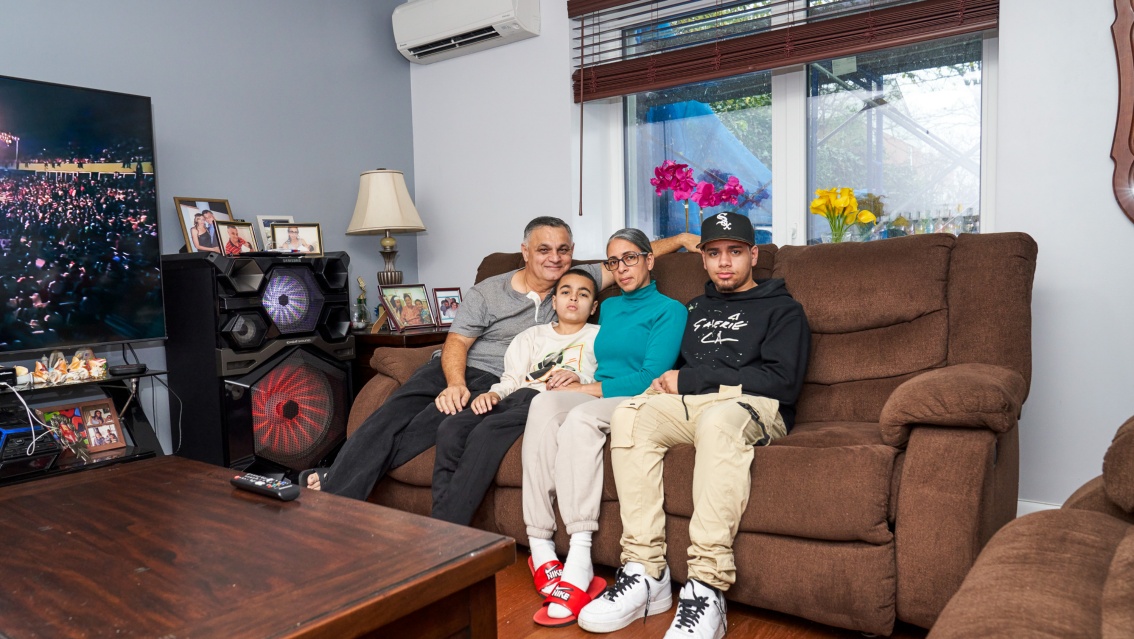 Eine vierköpfige Familie sitzt in ihrem Wohnzimmer auf der Couch, an der grauen Wand hängt ein großer Fernseher.